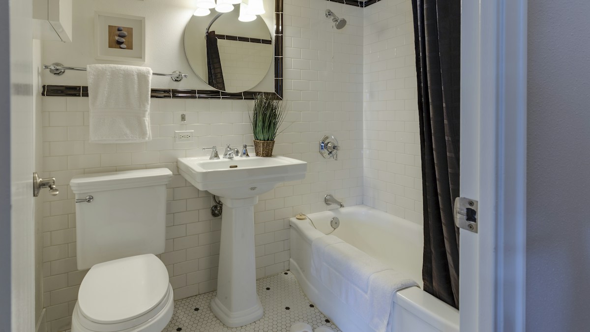 A white tiled bathroom where the toilet sits in the corner and the only electrical outlet is on the far wall beside the mirror, out of cord reach