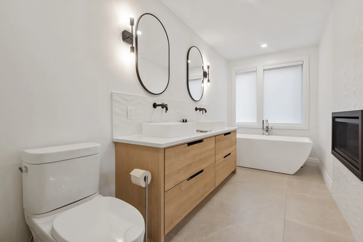 A bright modern bathroom with a toilet, oak vanity with dual oval mirrors, and a freestanding bathtub